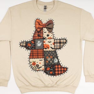 May include: Beige long-sleeved sweatshirt with a patchwork ghost design. The ghost is made of various patterned fabrics, including plaid, floral, and pumpkin prints. The ghost has a bow and is decorated with Halloween-themed elements.