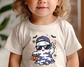 May include: Off-white t-shirt featuring a cartoon ghost wearing sunglasses and a blue cap, holding a jack-o'-lantern bucket. The ghost is giving a peace sign. Halloween-themed design with bats, stars, and spiderwebs.