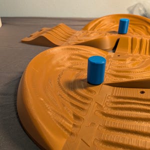 May include: A tan-colored, circular track with textured details and two blue cylindrical posts. The track has a raised, curved design, suggesting a race track or play set for toy cars.