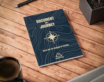 May include: A navy blue notebook with a topographic map design and the text "Document the Journey." A compass rose graphic is centered on the cover. The text "Miles are the measure of passion" is below the compass.