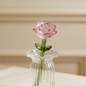 May include: A clear glass vase with a fluted design holds a delicate pink glass rose. The rose has detailed petals and a green stem with small leaves. The vase sits on a light-coloured surface, creating a soft, elegant aesthetic.