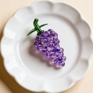 May include: A decorative glass grape cluster in shades of purple, with a green stem and leaves, rests on a white, scalloped-edge plate. The grapes are made of small, faceted beads. The plate sits on a light beige surface.