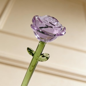 Puede incluir: Una rosa de cristal con una cabeza de flor lavanda y un tallo verde. La rosa tiene dos hojas verdes y está hecha de vidrio transparente. Los pétalos de la flor son detallados y el tallo es largo y delgado.