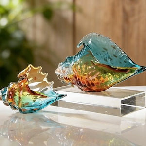 以下が含まれることがあります： 2つの装飾的なガラス製の貝殻。1つは左側に、もう1つは右側にあり、どちらも透明なベースが付いています。貝殻は青、オレンジ、黄色などのグラデーションカラーです。背景はぼやけており、木製の表面が見えます。