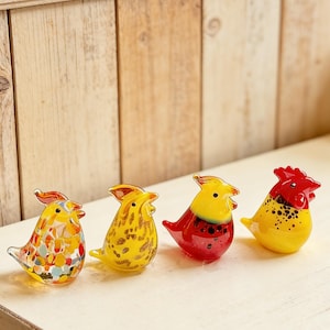 May include: Four colorful glass bird figurines. The birds are yellow, red, and clear with various patterns and designs. They are arranged on a white surface against a light wood background.