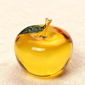 Puede incluir: Una manzana decorativa de cristal con cuerpo amarillo y hoja verde. El tallo es dorado con un toque de verde. La manzana está sobre una superficie beige.
