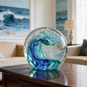May include: A clear glass paperweight with a wave design. The wave is depicted in shades of blue and teal, with bubbles throughout. The paperweight sits on a dark brown wooden table.