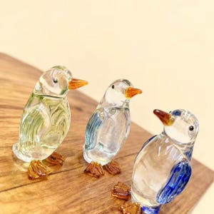 May include: Three glass penguin figurines with clear bodies, orange beaks, and amber-colored feet. One penguin has green accents, another has blue, and the third has blue wings. They are displayed on a wooden surface.