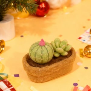 May include: A felt succulent arrangement featuring a green cactus with a pink top and a smaller green succulent in a brown felt pot. The arrangement is on a yellow surface with Christmas decorations.