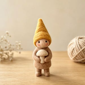 May include: A felt gnome figure with a yellow hat and a mushroom. The gnome is holding a white mushroom and has a light brown body. A ball of yarn and dried flowers are in the background.