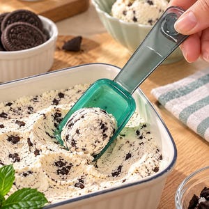 Könnte beinhalten: Ein durchsichtiger, dunkelgrüner Eisportionierer aus Kunststoff mit abgerundetem Griff und quadratischer Schaufel, der eine Kugel Cookies-and-Cream-Eis über einer weißen, rechteckigen Keramikschale hält, die mit demselben Eis gefüllt ist.