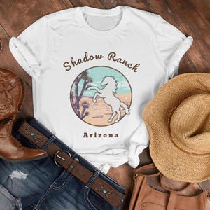 May include: White t-shirt featuring a circular graphic with a horse silhouette, desert landscape, and text "Shadow Ranch Arizona." The shirt is styled with a brown belt, denim jeans, a straw hat, and a brown leather bag.