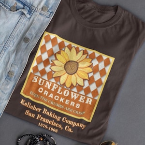 May include: A brown t-shirt featuring a sunflower design with the text "SUNFLOWER CRACKERS" and "Kelleher Baking Company San Francisco, Ca. 1878-1906". The shirt is made of a soft material and is suitable for casual wear.
