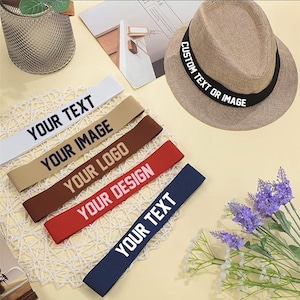 Puede incluir: Un sombrero fedora beige con una banda negra, junto a varias cintas de tela con opciones de texto personalizadas. Las cintas son blancas, beige, marrón, rojas y azul marino, cada una con texto blanco: "YOUR TEXT", "YOUR IMAGE", "YOUR LOGO" y "YOUR DESIGN".