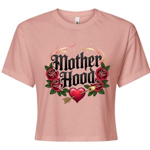 以下が含まれることがあります： ゴシック体の「Motherhood」の文字がプリントされた、明るいピンク色のクロップドTシャツ。赤いバラ、緑の葉、矢が刺さったハートで囲まれています。デザインはタトゥーにインスパイアされた美学を持っています。