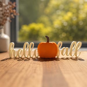 May include: A decorative sign featuring the white cursive text hello fall with an orange pumpkin replacing the letter o. The pumpkin has a brown stem and a smooth surface. The sign is constructed as a single piece with a flat base.