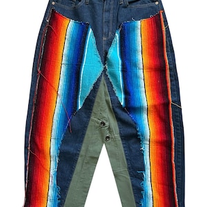 May include: Blue denim jeans with colorful woven fabric patches in red, orange, blue, and turquoise. The jeans have a unique design with green fabric panels and button details, creating a patchwork aesthetic.