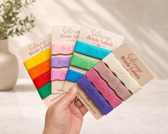May include: A collection of colorful silicone bottle labels attached to beige display cards. Each label features a wavy edge and is embossed with a custom name. The labels come in various colors including yellow, orange, red, green, pink, purple, blue, and cream, with the text Silicone Bottle Labels printed on each card.