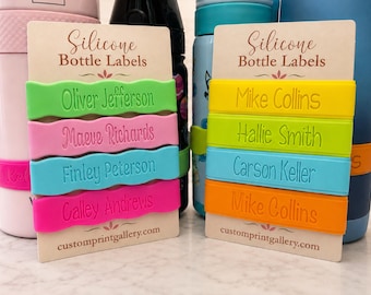 May include: Two rectangular display cards labeled Silicone Bottle Labels and customprintgallery.com, each holding four wavy silicone bands. The left card features green, pink, teal, and magenta bands with names. The right card features yellow, lime, blue, and orange bands with names.