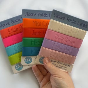 May include: A stack of colorful silicone bottle labels with personalized names, including "Elise A", "Mike Collins", "Justin M", "Mike C", "Fin", "Elizabeth M", "Callie Brown", "Elise Abel", and "Elizabeth". The labels are made by Custom Print Gallery.