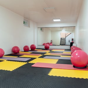 Pode incluir: Uma sala de fitness com um piso composto por peças de encaixe pretas e amarelas. Várias bolas de exercício cor-de-rosa estão colocadas sobre tapetes retangulares em tons de rosa e cinzento, dispostos por toda a sala.