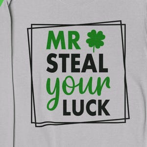Pode incluir: Um moletom cinza claro com o texto "MR STEAL your LUCK" em verde e preto, com um trevo. O moletom é cercado por decorações de trevo verde e um chapéu verde.