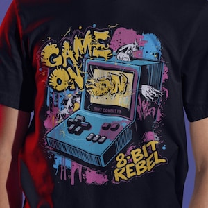 May include: A black t-shirt with a colorful graphic of a retro video game console and the words "GAME ON" and "8-BIT REBEL". The shirt is worn by a person wearing a headset with a microphone.
