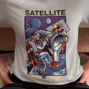 Pode incluir: Uma t-shirt cinzenta clara de manga curta com uma impressão gráfica de dois astronautas a realizar a manutenção de um grande satélite no espaço. O design inclui planetas, estrelas e o texto SATELLITE em letras amarelas a negrito.