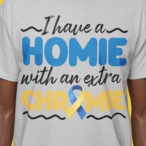Può includere: T-shirt grigio chiaro con la scritta "I have a HOMIE with an extra CHROME" in blu e giallo. La maglietta presenta un nastro blu e giallo ed è indossata da una persona.