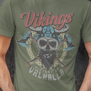Könnte beinhalten: Olivgrünes T-Shirt mit einem Wikinger-Schädel-Design, gekreuzten Äxten und dem Text "Vikings Victory or Valhalla". Die Grafik verwendet Blau, Rot und Gelb. Das Model trägt eine dunkelblaue Jeans.