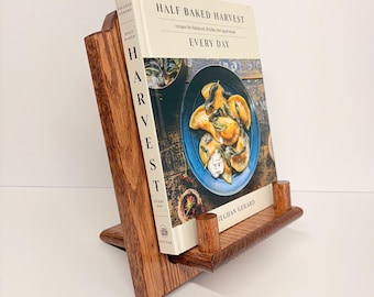 May include: A wooden cookbook stand with a book titled "Half Baked Harvest Every Day" is displayed. The stand is made of dark brown wood and has a slanted design to hold the book upright. The book cover features a blue plate of food.