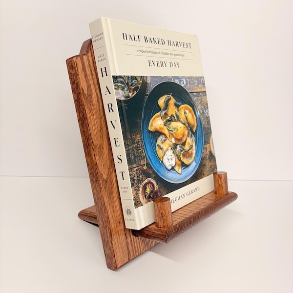 Pode incluir: Um suporte de livro de receitas de madeira com um livro intitulado "Half Baked Harvest Every Day" é exibido. O suporte é feito de madeira marrom escura e tem um design inclinado. A capa do livro apresenta um prato azul de comida.