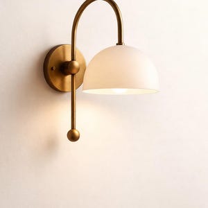 May include: A wall-mounted light fitting with a brass finish. It features a curved arm supporting a white, semi-spherical shade. The fitting is mounted on a round brass base.