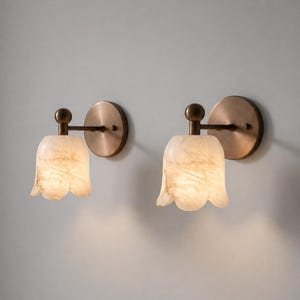 May include: Two wall sconces with a tulip-shaped, off-white alabaster shade. The sconces have a bronze-colored circular backplate and a matching arm and ball detail. The alabaster shades are illuminated, creating a warm glow.