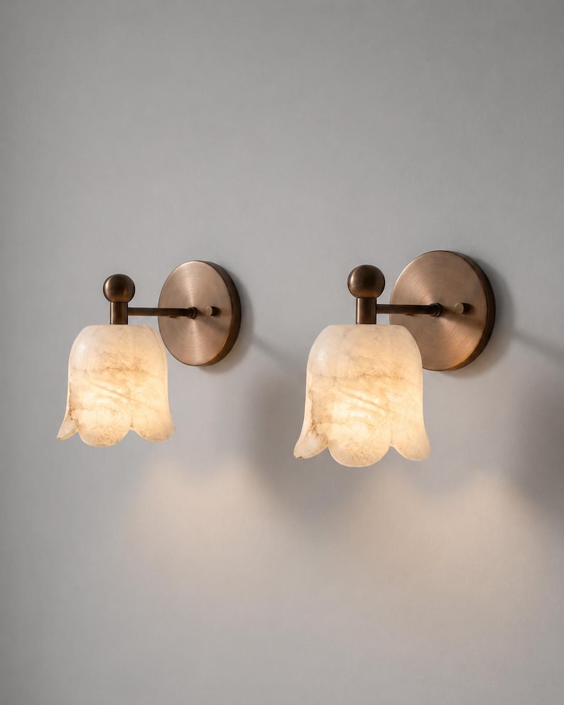 May include: Two wall sconces with a tulip-shaped, off-white alabaster shade. The sconces have a bronze-colored circular backplate and a matching arm and ball detail. The alabaster shades are illuminated, creating a warm glow.