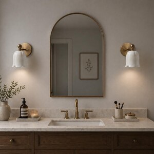 May include: A bathroom vanity with a marble countertop, a dark wood cabinet, and a gold-framed mirror. Two wall sconces with white tulip-shaped shades flank the mirror. A vase with greenery and a soap dispenser sit on the counter.