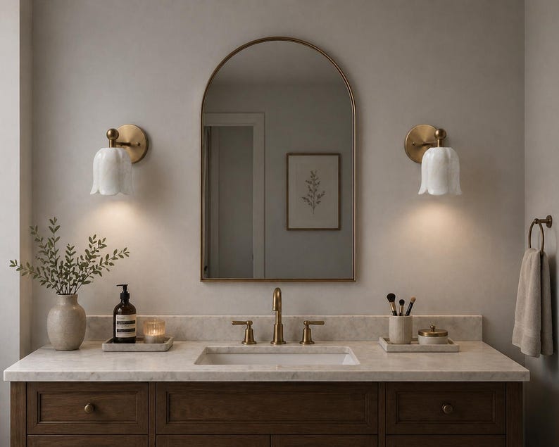 May include: A bathroom vanity with a marble countertop, a dark wood cabinet, and a gold-framed mirror. Two wall sconces with white tulip-shaped shades flank the mirror. A vase with greenery and a soap dispenser sit on the counter.