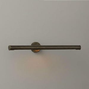 May include: A bronze-coloured, horizontal wall-mounted light fitting. The fitting has a circular base and a long, cylindrical bar with rounded ends. The light is mounted on a light grey wall.