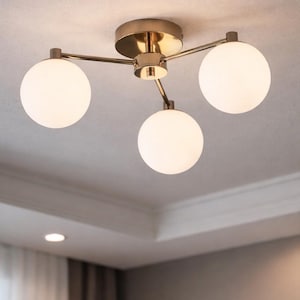 May include: A contemporary ceiling light with a brass finish. It has three spherical, frosted glass shades. The light is mounted on a white ceiling, and the fixture's arms extend from a central hub.