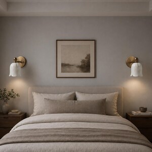 May include: A bedroom scene featuring a bed with neutral-toned bedding, a headboard, and a framed landscape painting. Two wall sconces with brass fixtures and white tulip-shaped shades flank the bed, providing soft lighting. Wooden nightstands are on either side.