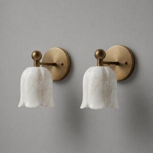 May include: Two wall sconces with a tulip-shaped, white marble shade and a brass-colored metal base. The sconces are mounted on a gray wall, providing a soft, ambient light.
