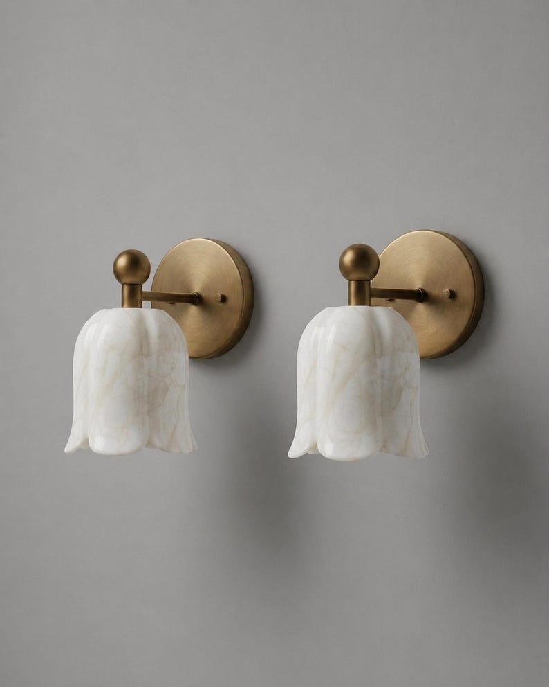May include: Two wall sconces with a tulip-shaped, white marble shade and a brass-colored metal base. The sconces are mounted on a gray wall, providing a soft, ambient light.