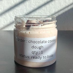 Puede incluir: Un tarro de cristal transparente lleno de masa de galletas de chocolate de color marrón claro. El tarro tiene una etiqueta blanca con el texto "order: chocolate cookie dough qty: 1 status: ready to bake". La masa tiene chispas de chocolate oscuro.