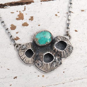 May include: A silver-toned necklace featuring a pendant with a turquoise stone and a cluster of textured, circular elements. The pendant is suspended from a delicate chain. The necklace is displayed on a white, distressed surface.