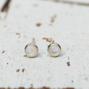 May include: A pair of round stud earrings featuring a smooth, iridescent white circular inlay set within a polished silver-toned metal bezel. The earrings have a simple post-back design, showcasing a clean, circular silhouette and a reflective surface.
