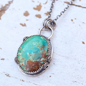 May include: A turquoise pendant necklace with a silver chain. The oval-shaped turquoise stone has a mix of blue and brown hues, set in a detailed silver bezel. The necklace is displayed on a white, textured surface.