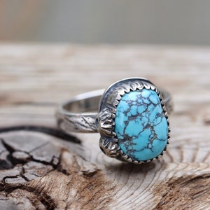 May include: A silver ring with a turquoise stone set in a detailed silver bezel. The stone has a blue-green color and a marbled pattern.