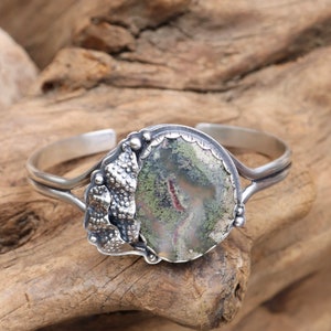 May include: A silver cuff bracelet with a green, brown, and white agate stone set in a silver bezel. The bezel is decorated with a silver design that resembles a cluster of leaves.