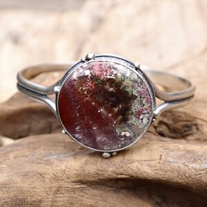 May include: A silver bracelet with a round, multi-colored agate stone set in the center. The stone has shades of brown, green, and white.
