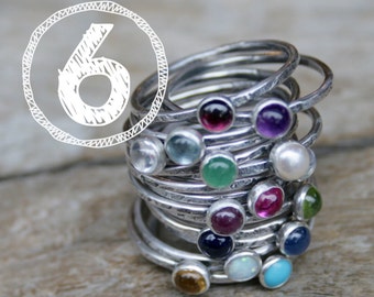 May include: A stack of sterling silver rings with various colorful gemstones, including amethyst, emerald, and turquoise. The rings are stacked on a wooden surface. The number "6" is in a white circle in the upper left corner.